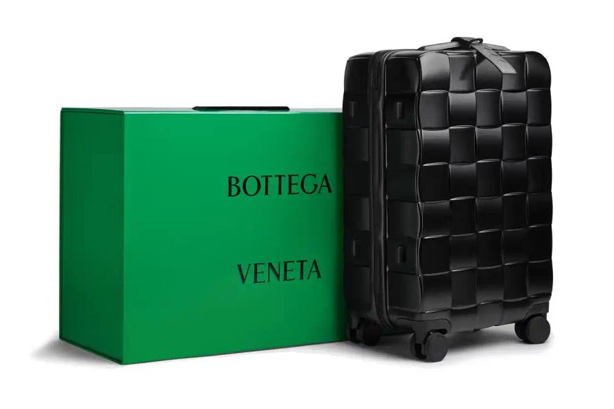 Bottega Veneta Odyssey carry-on suitcase with textured polycarbonate shell featuring 3D Intreccio pattern, silver hardware, and brown calfskin leather handles. The wheeled luggage is shown in a three-quarter view displaying its structured design and branded details.