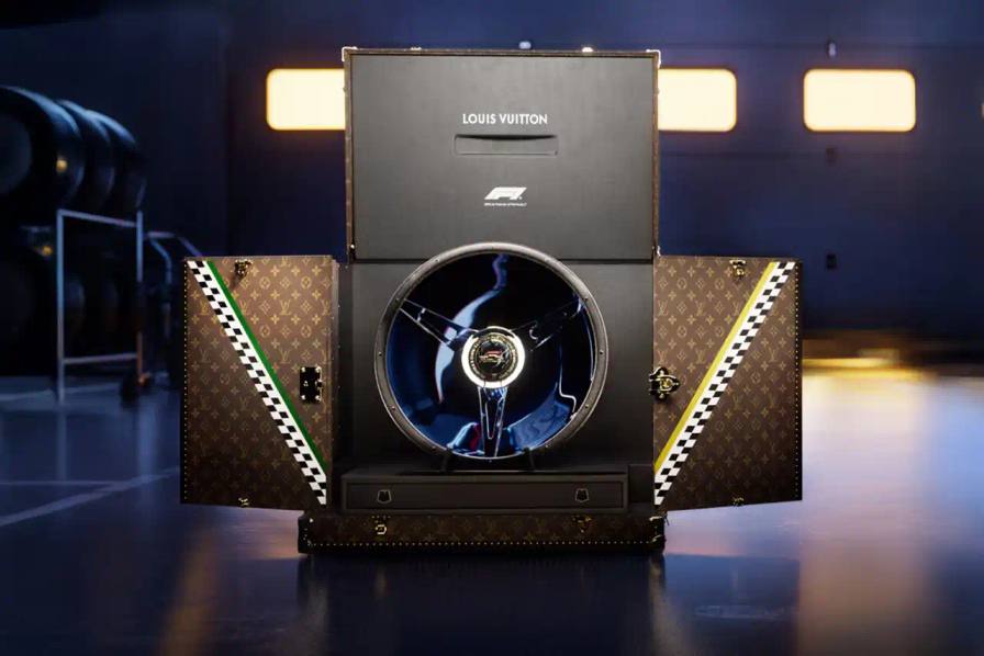 A Louis Vuitton trophy trunk with classic brown monogram pattern and gold accents. The trunk features diagonal green and gold checkerboard details on its doors, representing Australian national colors. When opened, it displays a Formula 1 steering wheel centerpiece. The trunk combines Louis Vuitton's luxury heritage with motorsport elements for the 2025 Australian Grand Prix.