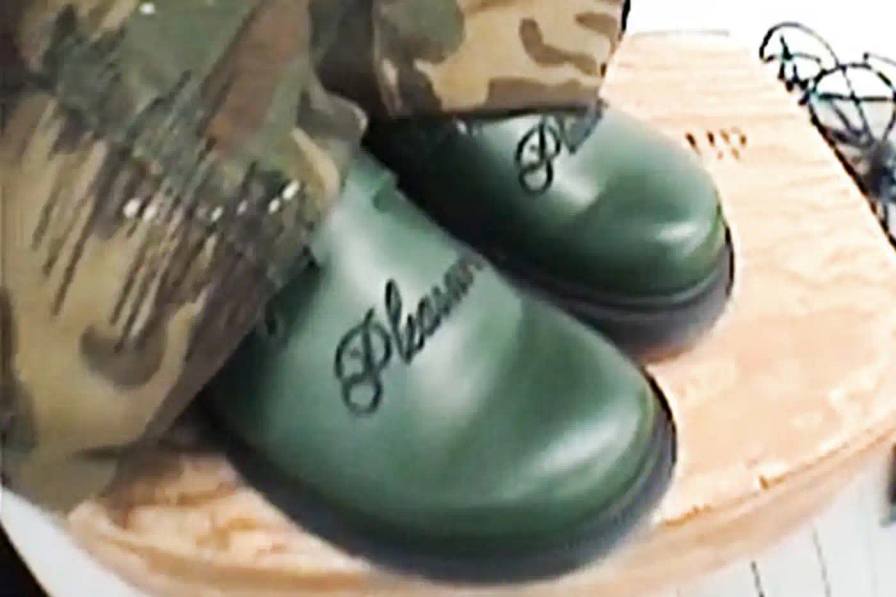 Green leather Dr. Martens Jorge Mule featuring black metal buckles, cursive PLEASURES branding stitched on the upper, signature yellow stitching, and a black Vibram outsole. The collaboration merges both brands' distinctive styles in a versatile slip-on design.