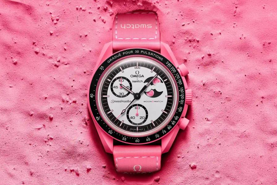 the new Mission to the Pink Moonphase MoonSwatch watch collaboration between OMEGA and Swatch. The timepiece features a pink Bioceramic case and strap, a moon phase complication with two pink moons, and a pulsometer scale for measuring heart rate. The article details the watch's design elements, including the heart motif on the push button, UV-light activated hidden details, and the cosmic-themed caseback. It also provides pricing ($310 USD), availability information (select Swatch stores starting April 1), and mentions this release follows the previous MoonSwatch 1965 model.