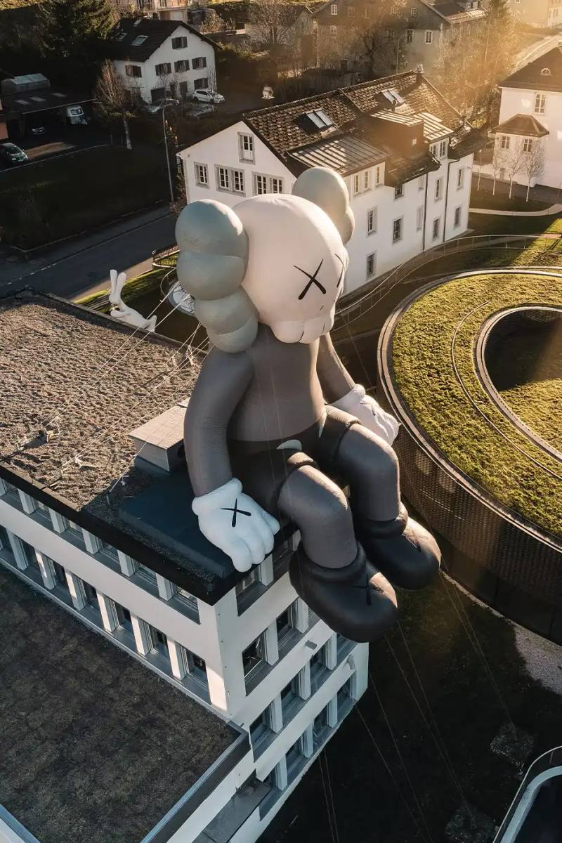 AllRightsReserved A massive 11-meter tall KAWS COMPANION sculpture in grey-toned color sits on the rooftop of the Audemars Piguet headquarters. The sculpture overlooks the glass spiral museum building in Le Brassus, Switzerland, against a mountainous backdrop, symbolizing the intersection of art and traditional watchmaking craftsmanship.