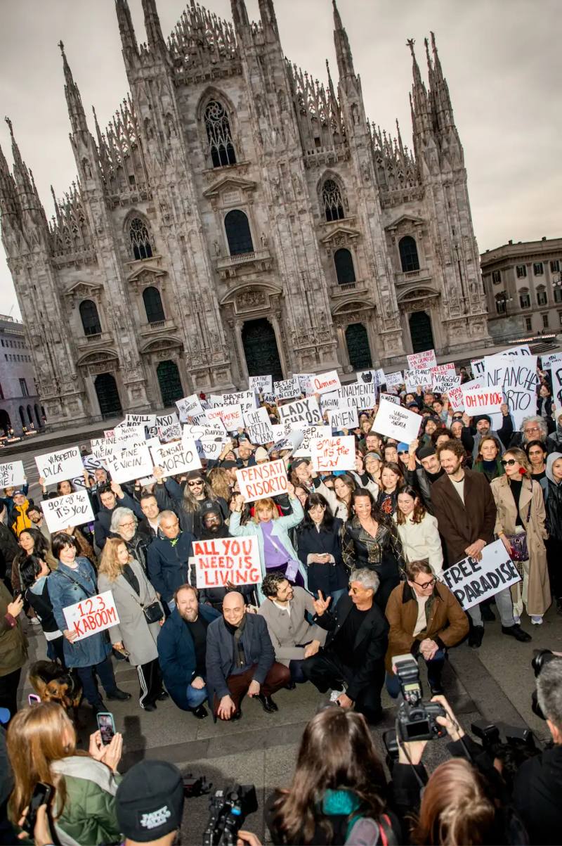 Photo of an art performance called "The Cheerful End" organized by artist Maurizio Cattelan and critic Nicolas Ballario in Milan. Held at dawn in Piazza Duomo, participants displayed humorous and philosophical signs about death. The event closed Milan Art Week and was supported by the Museo del Novecento. The performance used irony to encourage public reflection on mortality, with refreshments moved indoors at authorities' request. The article describes how the two creators acted as "accomplices in absurdity" to transform a somber subject into a thought-provoking shared experience.