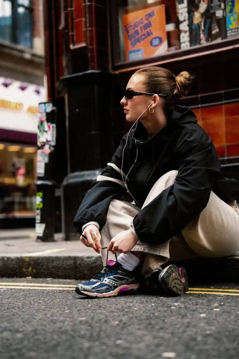 An image showing a person sitting on a city street wearing Saucony ProGrid Omni 9 sneakers in a dark colorway with pink accents.