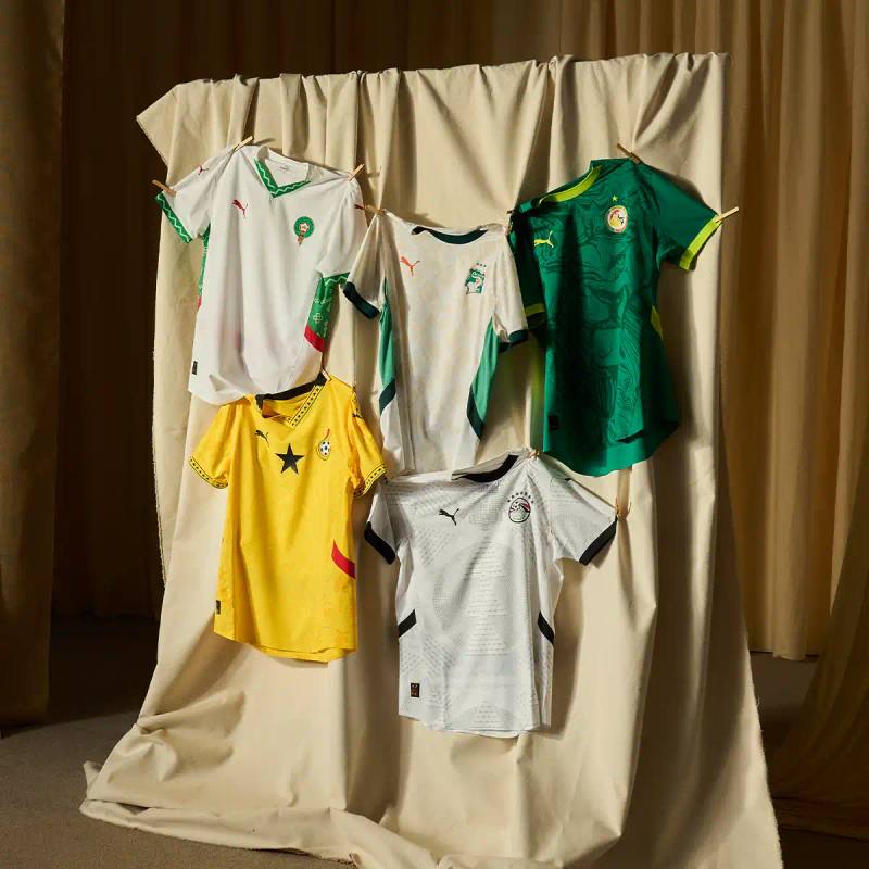 PUMA's 2025 AFCON collection displayed on mannequins, showing home and away jerseys for Morocco and Ghana national teams. The Morocco kit features geometric patterns in red and white, while Ghana's uniforms combine traditional symbols with yellow and white colorways.