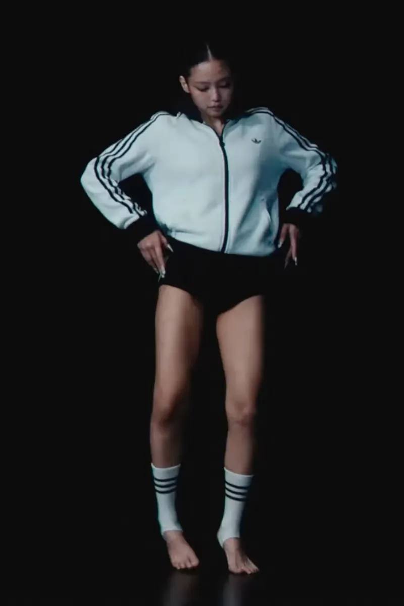 image of Jennie dancing against a black background wearing an Adidas tracksuit jacket, high-waisted shorts, and toe-less socks.