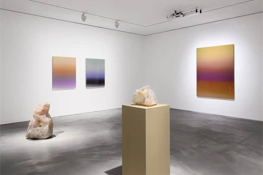 A gallery installation view featuring Mika Tajima's multidisciplinary artwork. Translucent acrylic paintings with gradient pigments, textile spectrograms of brain wave data, and rose quartz and glass sculptures that investigate technological mediation of human experience.