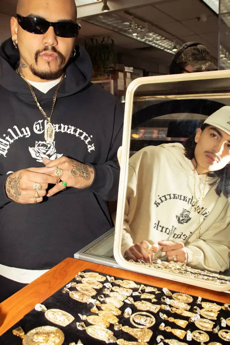 Image of Willy Chavarria's New York Collection featuring clothing items with Old English typography and the brand's signature rose logo. The collection includes hoodies, t-shirts, and caps in navy blue and off-white colors that showcase 'Willy Chavarria' arched above a rose emblem with 'New York' text below.