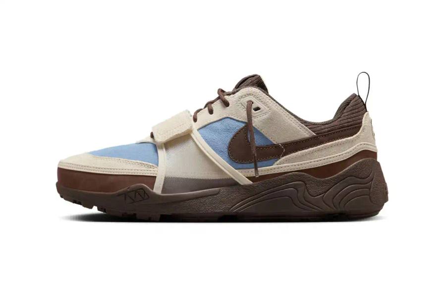 Image of the upcoming release of the Travis Scott x Nike Zoom Field Jaxx sneaker in "Leche Blue" colorway. It details the shoe's design inspiration from the Zoom Spiridon runner and Land Shark baseball cleat, describes the blue and brown color scheme, mentions the $170 price point, and notes the May 16 release date through Nike SNKRS and select retailers.