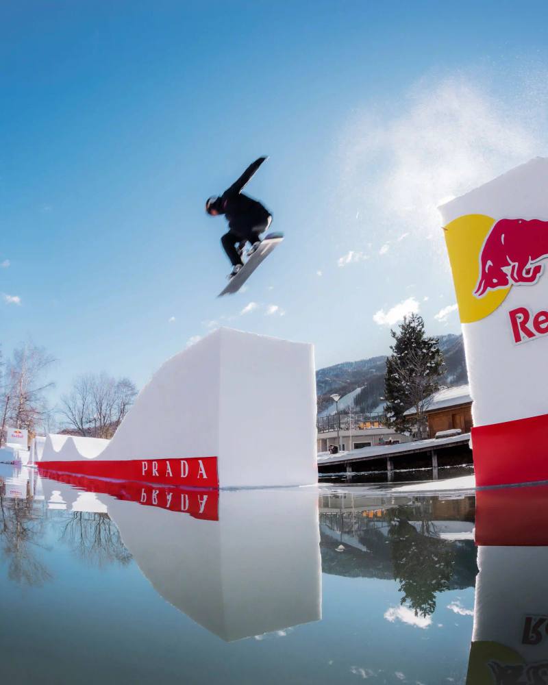 Snowboarder Pierre Vaultier riding on a narrow 50-centimeter track over a partially frozen lake in Serre-Chevalier. He wears black and red Prada Linea Rossa technical gear against a backdrop of snow-covered French Alps.