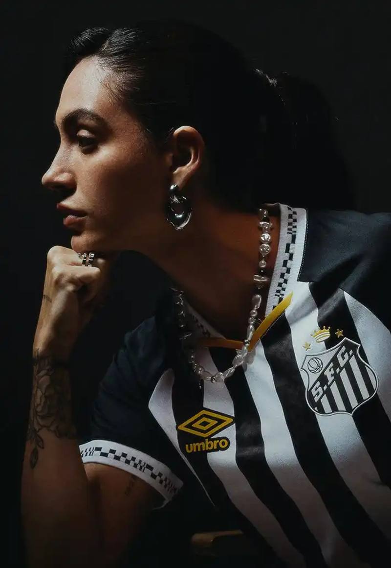 image of Santos FC's new 2025 away kit designed by Umbro. The kit features a black and white color scheme with gold accents on the Umbro logo, v-neck collar, and Santos crest. The design includes distinctive pixelated patterns on the collar and sleeve cuffs inspired by retro video games. The article mentions the kit's arrival midway through the season and notes its significance as the club welcomes back a key player wearing the number 10 jersey. The kit represents the passion of Santos supporters and is available for purchase through the official Santos FC online store.