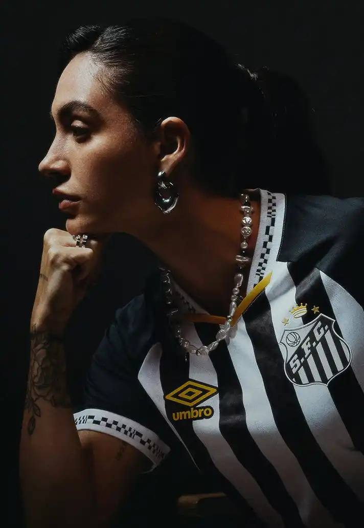 SANTOS FC UNVEILS 2025 AWAY KIT WITH UMBRO image of Santos FC's new 2025 away kit designed by Umbro. The kit features a black and white color scheme with gold accents on the Umbro logo, v-neck collar, and Santos crest. The design includes distinctive pixelated patterns on the collar and sleeve cuffs inspired by retro video games. The article mentions the kit's arrival midway through the season and notes its significance as the club welcomes back a key player wearing the number 10 jersey. The kit represents the passion of Santos supporters and is available for purchase through the official Santos FC online store.
