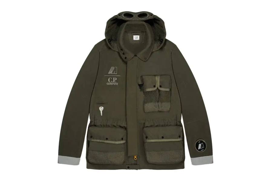 Automobili Amos The C.P. Company x Automobili Amos Mille Jacket shown in signature green color, featuring the distinctive goggle hood design, reflective details, and technical mesh construction. The jacket is photographed against a neutral background, displaying its motorsport-inspired details including the watch viewer window and specialized key pocket.