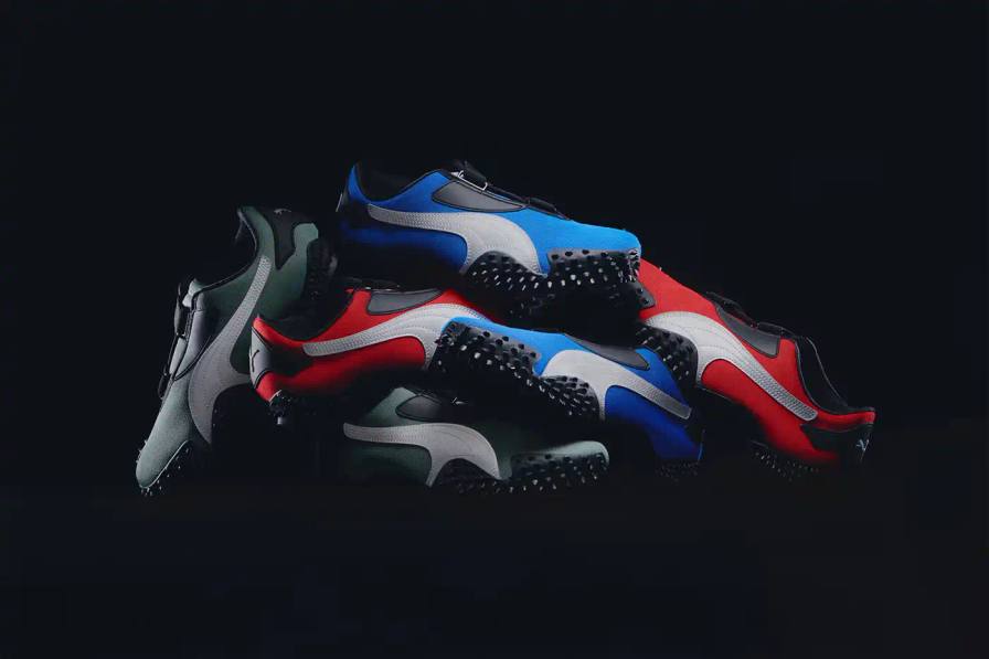 PUMA Mostro OG sneakers displayed in three colorways - Team Royal blue with silver accents, Green Moon green with silver detailing, and All Time Red with silver trim. Each shoe features the model's distinctive spiked sole, mesh upper, and hook-and-loop fastening system.