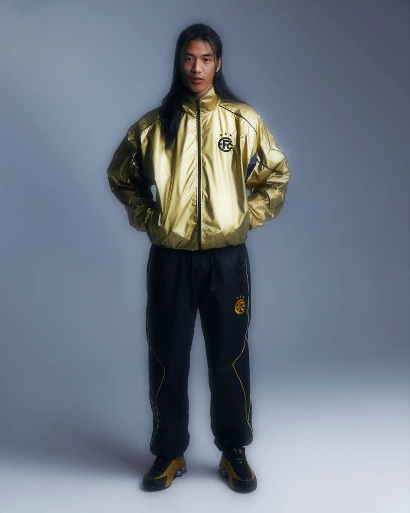 Image shows Equipo FC's reversible "Winners Tracksuit" displayed from both sides - one completely gold and one black with gold details. The tracksuit features star patterns and an embroidered crest, inspired by iconic 2000s football team apparel.