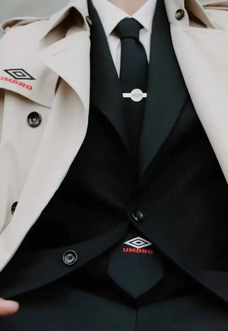 photo of fashion collaboration between sportswear brand Umbro and Italian label LC23 for Spring/Summer 2025. The collection draws inspiration from 1990s football manager attire, featuring items such as a leather Speciali sneaker with interchangeable tongue, a beige trench coat, a knitted football shirt, and accessories including a blazer, shorts, tie, and cap. The collection blends formal touchline style with contemporary streetwear and will be available from April 23 on lc23.it.