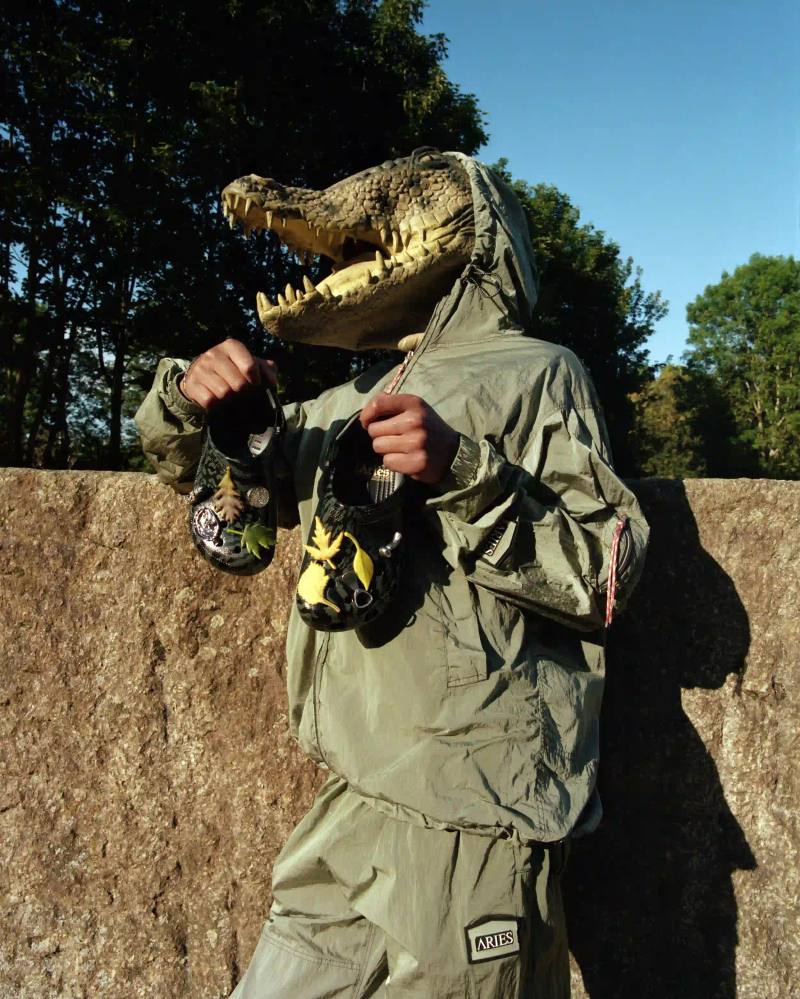 image about the second collaboration between London streetwear brand Aries and footwear company Crocs. The collection features two camouflage-patterned Classic Clogs in High Shine Black and Dark Algae Green colorways, accompanied by custom Jibbitz charms inspired by urban artifacts. The products launch July 23, 2025, priced at £75.00, available through Aries' website, flagship store, Crocs app, and select retailers worldwide.