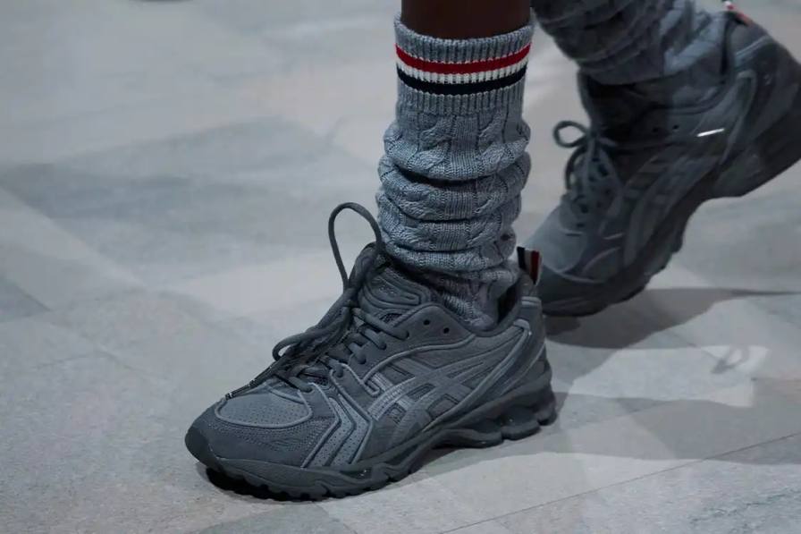 Thom Browne partnered with ASICS to release a redesigned GEL-KAYANO 14 sneaker featuring grey suede, mesh construction, and the designer's tri-color flag detail. The collection launches globally on March 2, 2026.