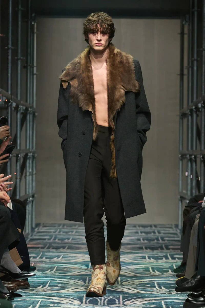 A fashion show staged within a three-story industrial metal pipe structure at Fondazione Prada. The runway features male models walking through narrow corridors under shifting blue, red, and yellow lights. The industrial set is softened by a luxurious carpet. Models wear oversized coats, exposed chests with fur stoles, cottage-print tops, plaid trenches, and Western-influenced footwear. The collection demonstrates a mix of formal and casual elements, with unexpected combinations like floral brooches on tailored blazers. The space combines elements of both a nightclub and a traditional ballroom setting.