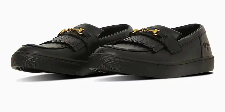 Converse All-Star Coupe GB Loafer shown from side angle, featuring minimalist leather construction, tasseled midfoot strap with embossed star details, and classic Converse sole. Available in black and off-white colorways.