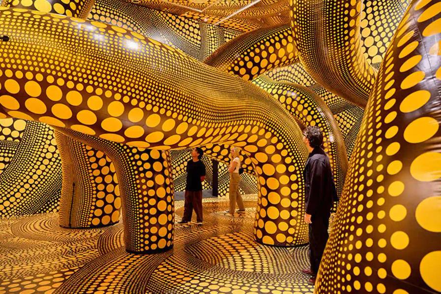 A photograph of Yayoi Kusama's immersive infinity mirrored room, featuring reflective surfaces creating a kaleidoscopic environment of endless light and geometric patterns. Polka-dotted sculptures and vibrant yellow and black elements are visible, representing the artist's signature visual style.