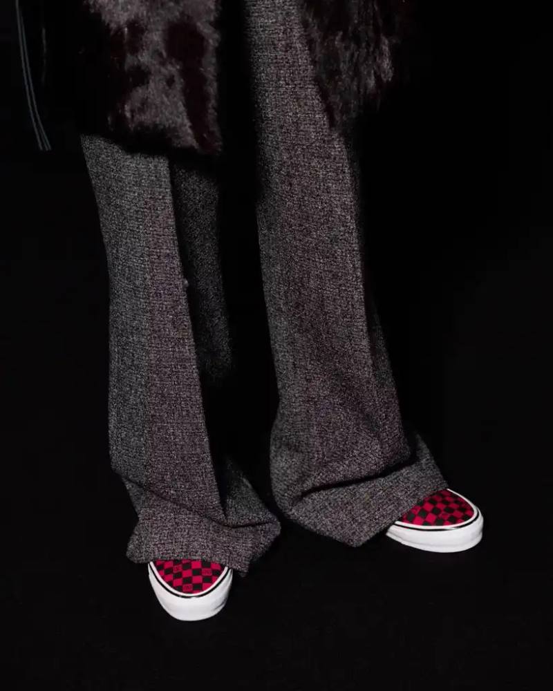 Image shows Valentino x Vans collaboration sneakers featuring checkerboard patterns with Valentino's "V" logo in white, red, pink, and black color schemes, displayed on runway models during Paris Fashion Week in a public bathroom-themed set.