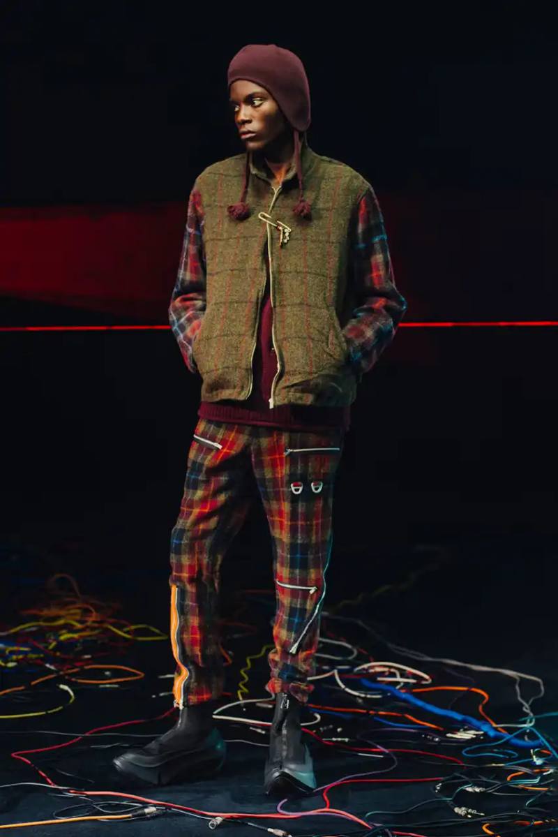 Fashion runway image featuring models wearing black tailoring and graphic prints, with neon lighting and monster illustrations by Josep Baqué, representing UNDERCOVER's FW25 collection exploring punk and musical subcultures.
