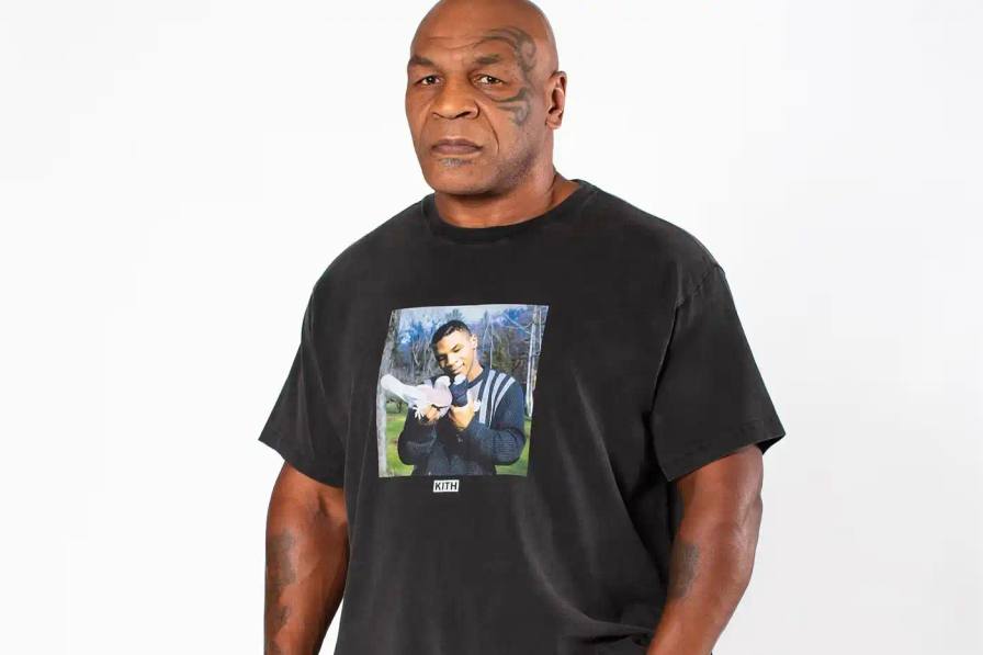 The image shows Mike Tyson, the famous boxer, gently pressing two pigeons together as if they are kissing. This is a recreation of the "Now Kith" meme, which references Tyson's distinctive lisp. Tyson is wearing one of the collaborative t-shirts from his Kith collection, which features the meme image in a vintage-inspired design.