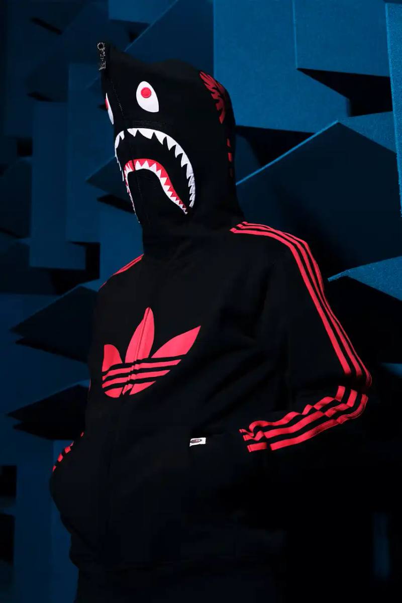 This article discusses the new collaboration between adidas and A Bathing Ape (BAPE). It details their latest capsule collection centered around the SSTR V BAPE sneaker, which reimagines the classic Superstar silhouette. The article describes the sneaker's design features, available colorways for adults and children, and mentions the accompanying apparel and accessories including tracksuits, hoodies, t-shirts, caps, pouches, and socks. It concludes with information about the April 5th release date through the CONFIRMED app and select retailers.