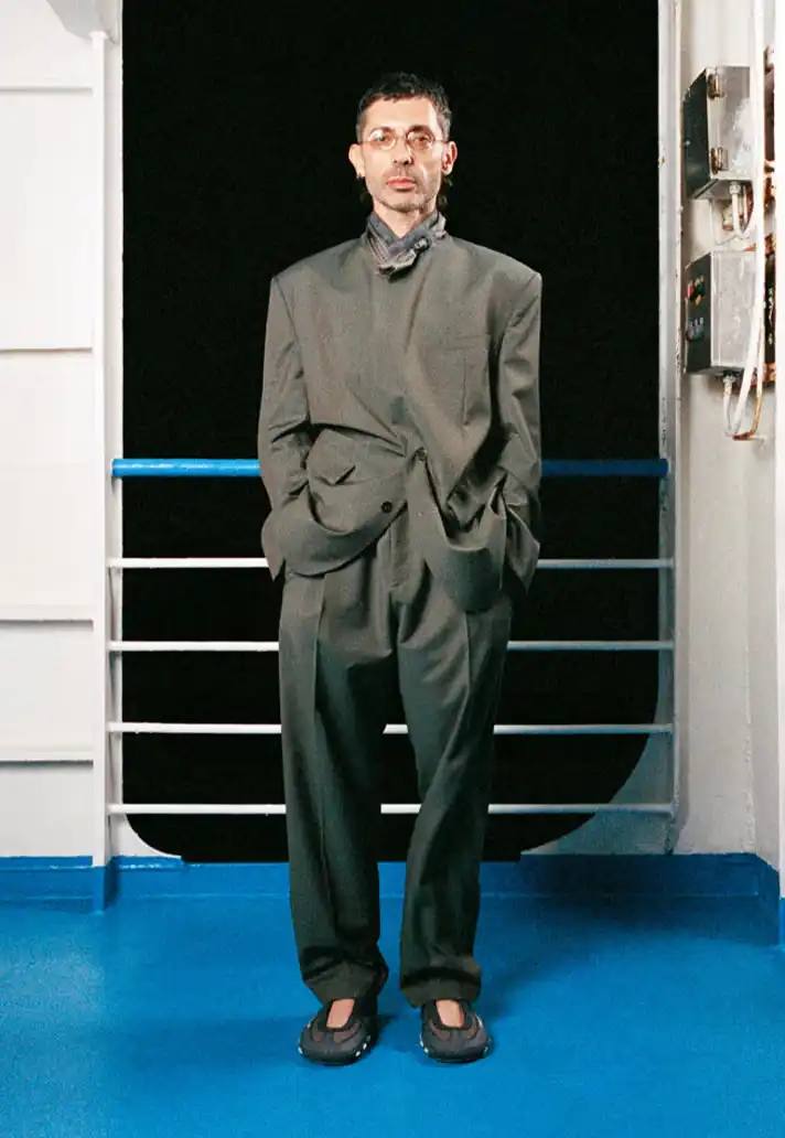 MAGLIANO SHOWS SS26 COLLECTION VIA FERRY FILM image about Italian designer Luca Magliano's Spring/Summer 2026 menswear collection, presented as a film aboard a ferry during Milan Fashion Week. The collection features camping and nomadic influences, with repurposed materials, loose silhouettes, and collaborative footwear including Beja soccer sneakers and 1900s-inspired boots. Shoot by Leonardo Scotti.