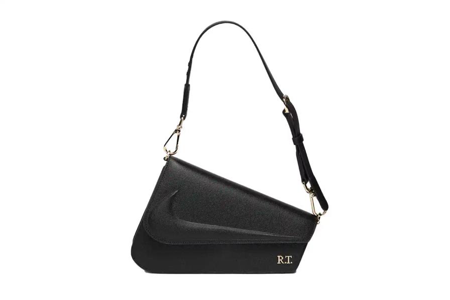 The Mercer Bag by Nike x Riccardo Tisci, shown in black pebbled leather with gold hardware. The minimalist design features a front flap with debossed Nike logo and R.T. metal signature at the corner. The bag includes an adjustable shoulder strap.