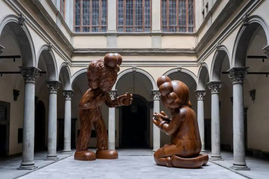 KAWS AT PALAZZO STROZZI IN FLORENCE KAWS installs large wooden sculptures at Florence's Palazzo Strozzi, featuring his signature characters in poses referencing Fra Angelico's 1433 Annunciation painting, exploring digital age themes through January 26, 2025.