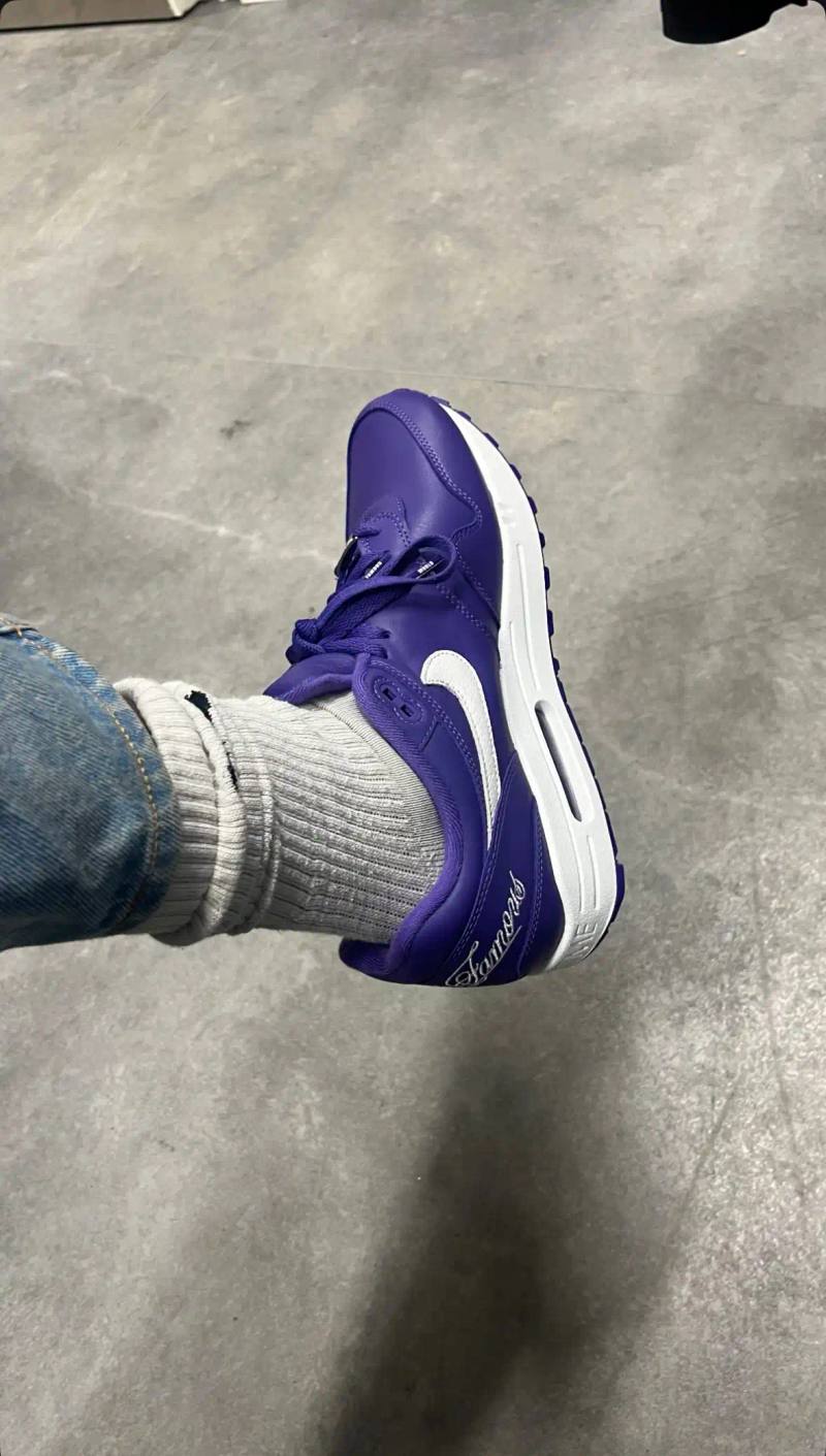Close-up photograph of the Supreme x Nike Air Max 1 '87 in Varsity Purple colorway showing white Supreme "World Famous" script on the purple heel counter, white midsole with debossed SUPREME wordmark, and purple upper with white Nike swoosh logo on the side.
