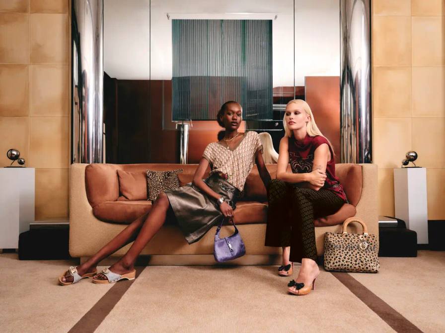 Two women sit on a sofa wearing designer clothing and accessories including leather skirts and animal print bags in a modern interior setting.