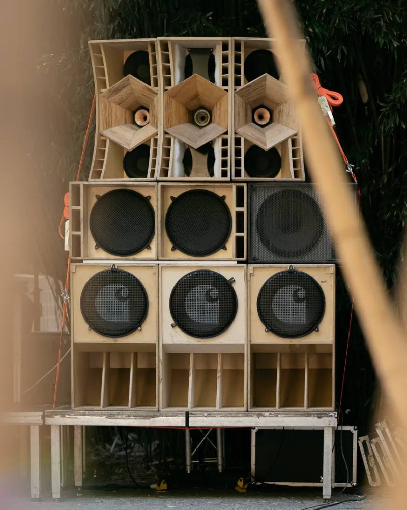 A stacked wall of custom wooden speakers sits outdoors, featuring the Carhartt C logo on the grilles held together by orange straps. image by Stefano Mattea