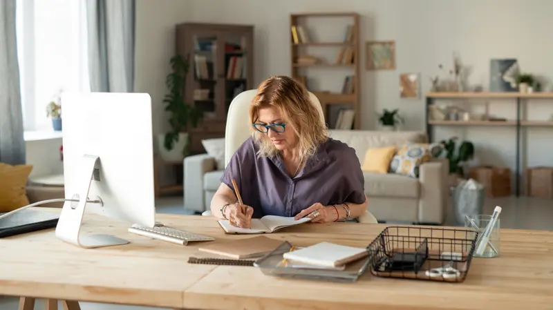 A woman working at a desk with a desktop computer in a bright, open home office with bookshelves and natural light.