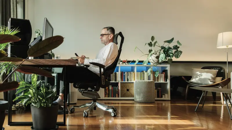 A man working at a desk in a well-appointed home office surrounded by plants and natural light.A man working at a desk in a well-appointed home office surrounded by plants and natural light.