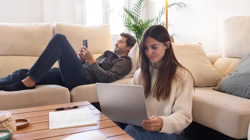 A woman using a laptop on the floor of a bright living room while her partner relaxes on the couch with a phone.