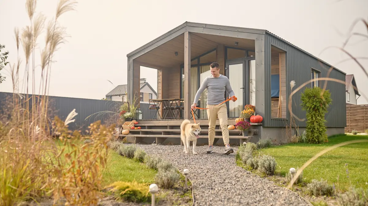 A man walking his dog along a garden path in front of a modern detached guest house on a residential property.