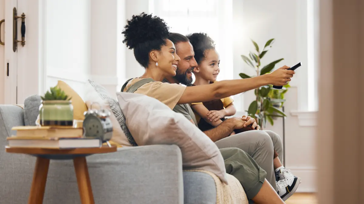 A family sitting together on the couch watching TV in a bright, modern living room, with the mom pointing the remote at the screen.
