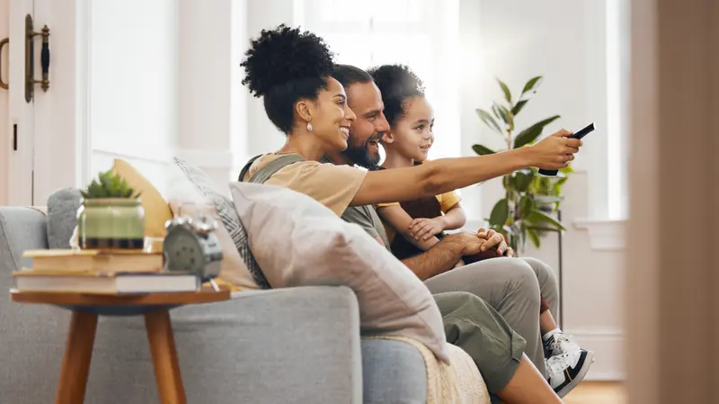 A family sitting together on the couch watching TV in a bright, modern living room, with the mom pointing the remote at the screen.