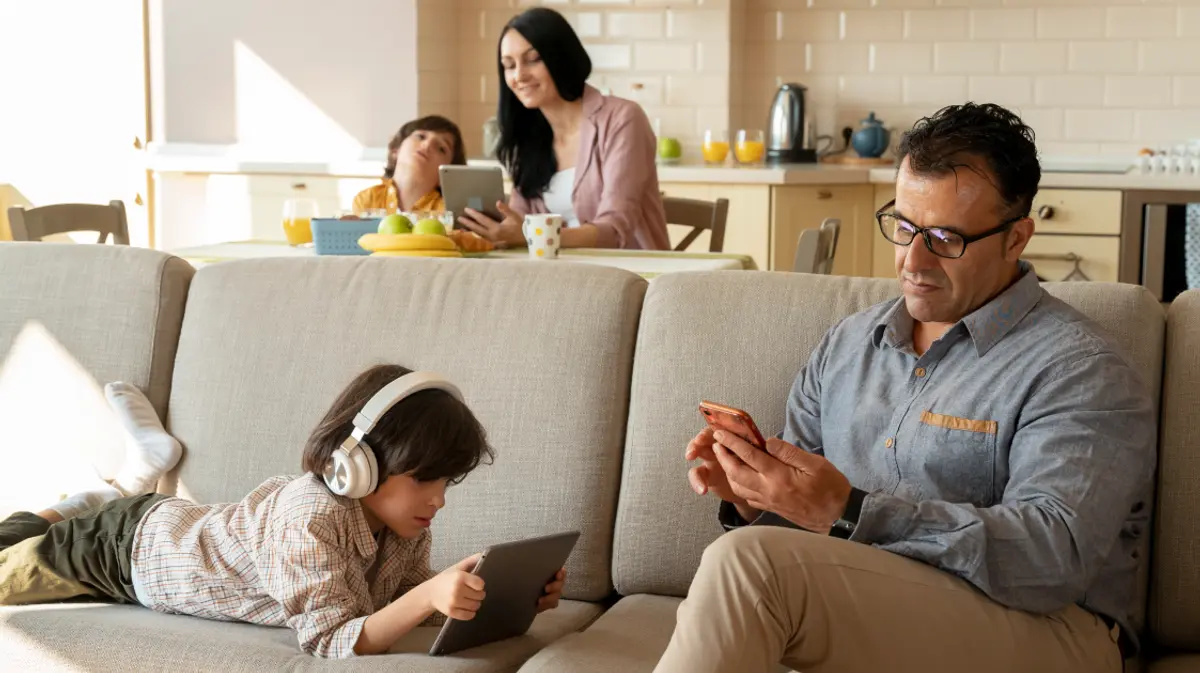 A family at home with multiple devices in use simultaneously, a child on a tablet with headphones on the couch, a father on his phone, and a mother and child with a tablet in the kitchen background.