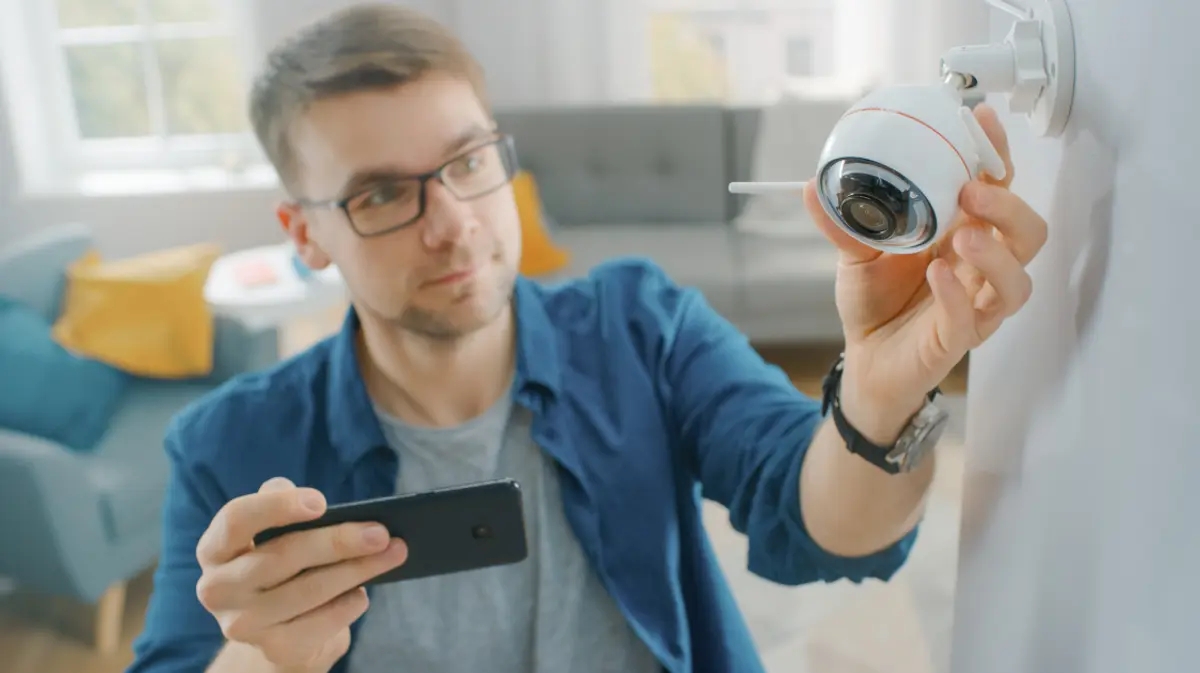 A man adjusting a Wi-Fi security camera mounted on a wall while checking the setup on his smartphone in a modern living room.