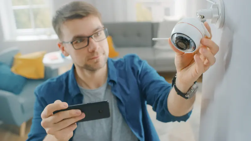 A man adjusting a Wi-Fi security camera mounted on a wall while checking the setup on his smartphone in a modern living room.