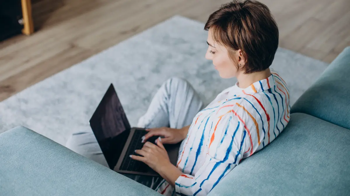 A woman working on a laptop from the couch in a bright, modern living room.