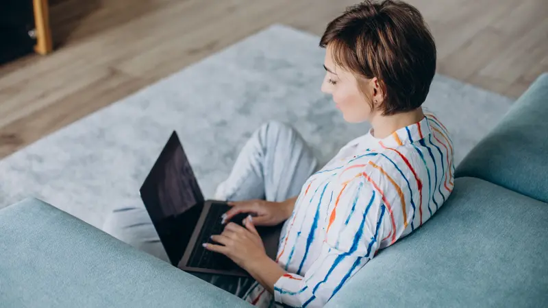 A woman working on a laptop from the couch in a bright, modern living room.