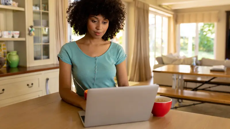 A woman working on a laptop at her kitchen table in a bright, open-plan home.
