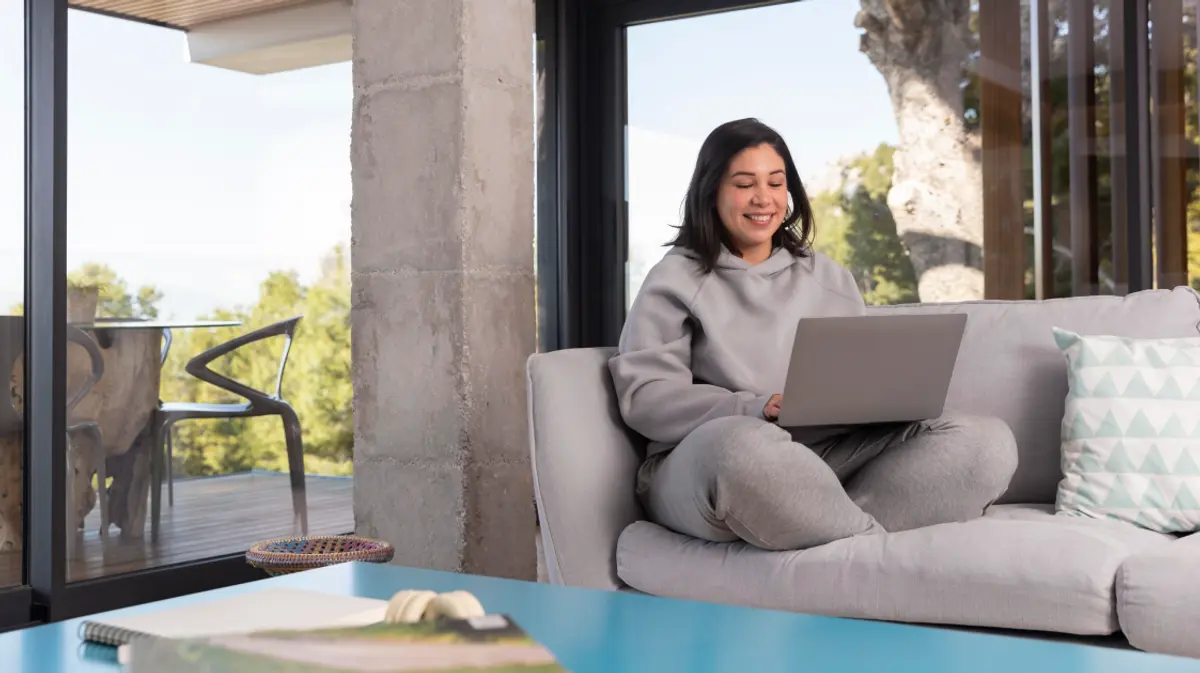 Woman relaxing on a sofa with a laptop in a spacious modern home with floor-to-ceiling windows and an outdoor view