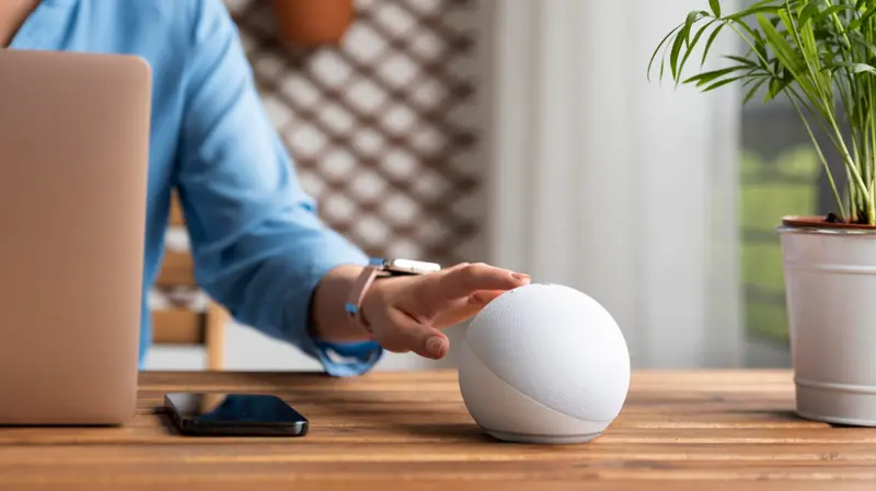 Person reaching to tap a smart speaker on a home office desk, with a laptop and phone nearby