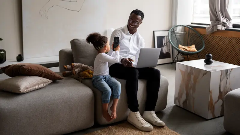 A father working on a laptop on the couch while his young child shows him something on a phone in a modern living room.