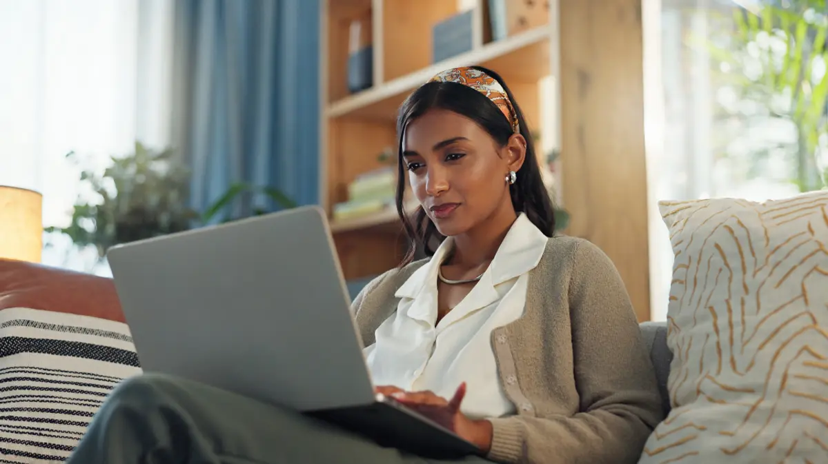 A woman working on a laptop on the couch in a warm, well-decorated living room with natural light and bookshelves in the background.